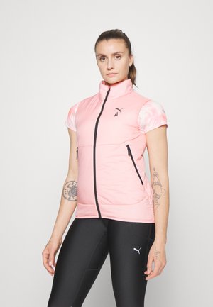 Giacca senza maniche rosa con colletto alto, chiusura a zip, due tasche laterali e dettagli neri. Indossata sopra una camicia a maniche corte a fantasia.