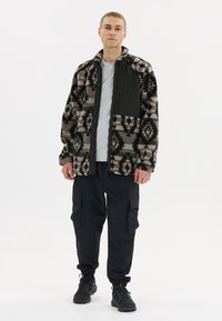 Fleecejacke mit schwarzem geometrischem Muster und solidem schwarzen Einsatz; graues Hemd darunter. Schwarze Cargo-Hosen und Schnürstiefel.
