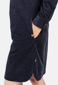 Granatowa sukienka z korduroy o luźnym kroju, z bocznymi kieszeniami i zaokrąglonym dołem. Posiada małą metkę z logo przy kieszeni w celach identyfikacyjnych.