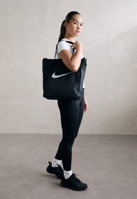 Jonge vrouw in zwarte leggings en een witte top, met een grote zwarte Nike-tas over haar schouder, staande op een grijze vloer.