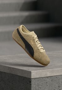 Μπεζ αθλητικό παπούτσι με μαύρη ρίγα και μικρά μαύρα πουά, δεμένο με κορδόνια, ακουμπισμένο σε γκρίζα τσιμεντένια επιφάνεια με σκαλοπάτια στο φόντο.