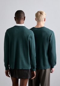 Two individuals wearing long-sleeve, ribbed sweaters in teal. One sweater has a collar, the other a rounded neck. Simple, textured fabric.