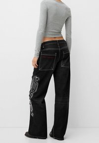 Mustat farkut, joissa on leveät lahkeet ja kuluneet yksityiskohdat sivuilla sekä punainen "new runner" -kirjonta. Mukana on kaksi takataskua, joissa on kontrastivärinen ompelu.