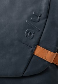 Dark blue fabric bag detail with embossed "alu" text and stitched brown leather strap.