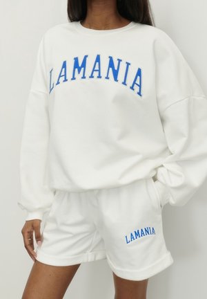 Persoon draagt een witte trainingspak met blauwe "LAMANIA" tekst op de borst en linker dij, poseert met één hand in de zak.