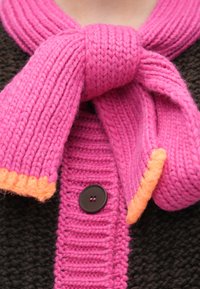 Pletený kardigán s tmavo hnedým telom, s jasnoružovou šálou uviazanou na krku a oranžovými akcentmi na okrajoch a gombíku.