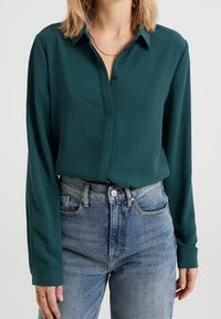 Grön långärmad blus med knäppning fram, krage och slät textur, i kombination med högmidjade blå jeans.