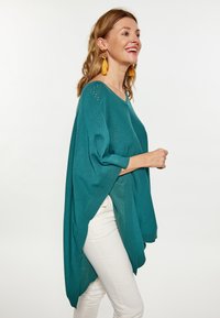 Poncho tricotat în nuanța teal, cu mâneci scurte tip batwing, decorat cu modele lucrate în dantelă și tiv elastic, asortat cu pantaloni albi.