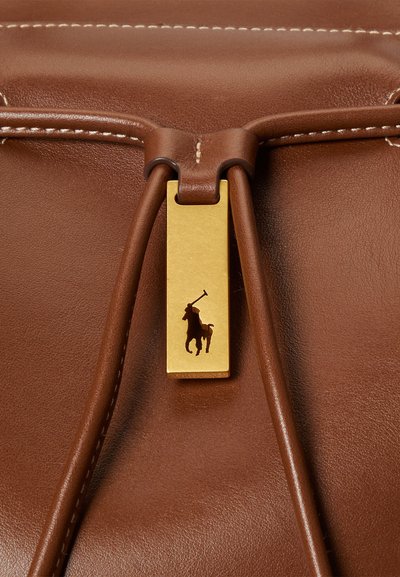 Λεπτομέρεια καφέ δερμάτινης τσάντας με χρυσό ορθογώνιο κούμπωμα που φέρει μαύρη σιλουέτα καβαλάρη, στερεωμένο με δερμάτινα κορδόνια.