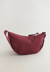 Mørkerød stof crossover taske med en buet form, lynlåslukning og dobbelte sideløsninger for nem transport. Glat tekstur med minimalistisk design.