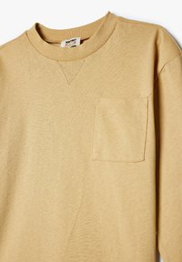 Beige långärmad skjorta gjord av texturerat bomull, med rundad halsringning och en enda bröstficka med sömnadsdetaljer.