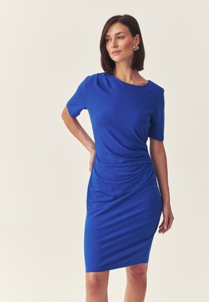 Kobieta o brązowych włosach do ramion, ubrana w złote kolczyki i dopasowaną sukienkę w królewskim błękicie, stojąca z jedną ręką na biodrze na jasnym tle.