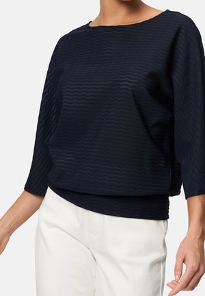 Frau trägt ein dunkelblau strukturiertes Langarmshirt mit rundem Ausschnitt und weiße Hose, abgebildet vom Hals bis zur Hüfte.