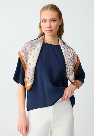 Femme aux cheveux blonds portant un chemisier bleu marine, un pantalon blanc et une écharpe blanche avec des motifs cachemire bordeaux et des bords marron, drapée sur ses épaules.