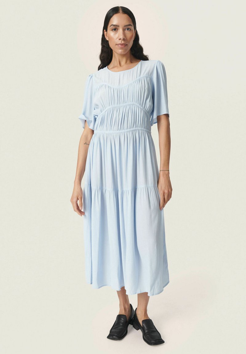Rochie midi de un albastru deschis, cu un bust plisat, mâneci scurte tip bulă și fustă în straturi. Material neted cu o croială lejeră. Pantofi negri, tip slip-on.