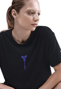 Kvinde med tilbageholdt hår iført en sort T-shirt med et lilla FC Barcelona-logo og tekst, der ser mod sin højre side mod en hvid baggrund.