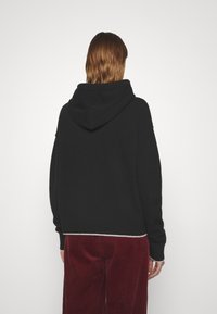 Sweat à capuche noir, oversize, avec une grande capuche, des poignets et un ourlet côtelés, et une bordure blanche contrastante en bas. Porté avec un pantalon en velours côtelé bordeaux.