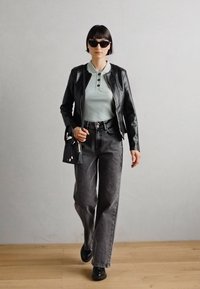 Giacca di pelle nera sopra un maglione a coste verde chiaro, jeans grigi a gamba larga e scarpe nere, con occhiali da sole neri e una piccola borsa.