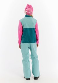 Fleecejack in teal en roze met een hoge kraag, gecombineerd met aqua skibroek. Het model draagt een roze en witte gestreepte gebreide muts.