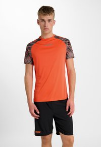 Orange idrottströja med svarta mönstrade ärmar, med rund hals och reflekterande logotyp. Matchas med svarta shorts med lättviktsdesign.