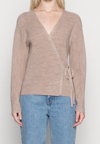 Beige stickad omslagströja med V-ringning, långa ärmar och knytning på sidan. Texturen är ribbad, kombinerad med ljusblå jeans.