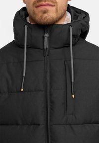 Mann trägt eine schwarze gepolsterte Jacke mit hochgeschlossenem Kragen und Kapuze, die schwarze und weiße Zugbänder mit gelben Spitzen aufweist.