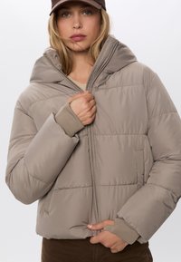 Žena u taupe puffer jakni i smešnim šeširom, zatvara jaknu s obje ruke, na jednobojnoj svijetloj pozadini.