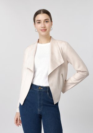 Joven mujer que lleva una chaqueta beige abierta por delante, camisa blanca y vaqueros azules, de pie con una mano en la cadera sobre un fondo liso.