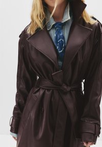 Trench coat in ecopelle bordeaux con ampi risvolti, vita con cintura, tasche laterali e camicia in denim a contrasto con una sciarpa blu.