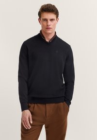 Mann trägt einen schwarzen V-Ausschnitt-Pullover über einem braunen Hemd, kombiniert mit braunen Hosen, vor einem einfarbigen hellen Hintergrund stehend.