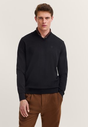 Mann trägt einen schwarzen V-Ausschnitt-Pullover über einem braunen Hemd, kombiniert mit braunen Hosen, vor einem einfarbigen hellen Hintergrund stehend.
