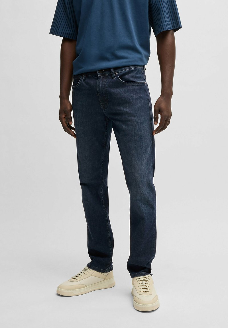 Mörkblå denimjeans med rak passform, som har subtila blekningsdetaljer, klassisk femficksdesign och dragkedja. Bärs med beige sneakers.