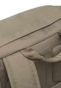 Beigen värinen kangasreppu, jossa on teksturoitu pinta, yläkahva, pehmustetut olkahihnat ja vetoketjullinen tasku.