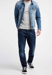 Giacca in denim con lavaggio azzurro chiaro, maglione grigio, jeans blu scuro e sneakers bianche e nere, che mostrano un look casual a strati.