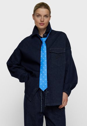 Frau trägt eine locker sitzende dunkle Jeansjacke und Hose mit elastischen Bündchen sowie eine hellblaue gemusterte Krawatte und steht mit einer Hand in der Hüfte.