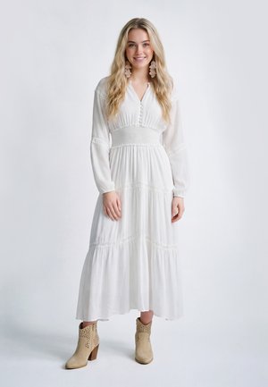 Giovane donna con lunghi capelli biondi che indossa un vestito maxi bianco a maniche lunghe e stivaletti beige, in piedi e sorridente su sfondo bianco.