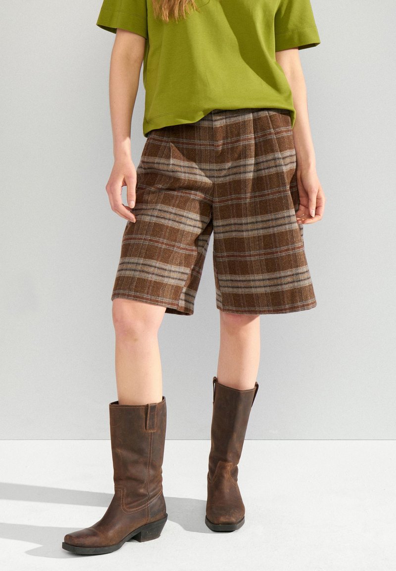Personne portant un short marron à carreaux jusqu'aux genoux, un haut vert à manches courtes et des bottes en cuir marron foncé mi-mollet, debout sur un sol clair.