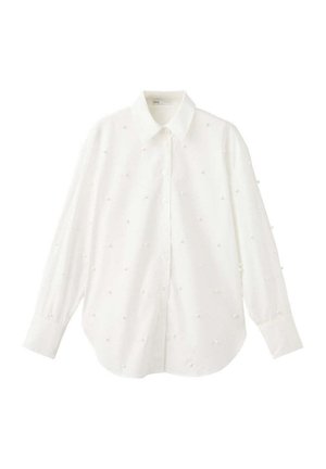 Femme portant un chemisier blanc à manches longues et une jupe midi blanche texturée, tenant une pochette noire, et portant des sandales noires à talons.