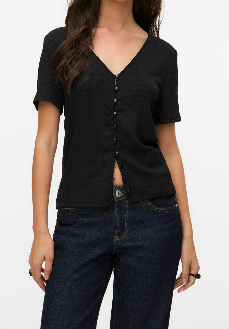 Femme portant un haut noir à manches courtes en V avec des boutons sur le devant et un jean bleu foncé sur un fond uni.