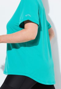 Osoba běžící v tyrkysovém sportovním tričku s krátkým rukávem a černých legínách na bílém pozadí.