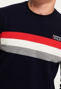 Námořnicky modrý svetr s vodorovnými pruhy v červené, šedé a bílé barvě. Žebrovaný kulatý výstřih, hebká textura, s logem na horní levé straně.