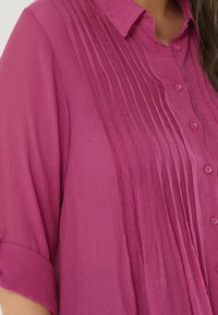 Camisa fucsia con cierre de botones en la parte delantera, con pliegues verticales y mangas enrolladas. Fabricada en un tejido ligero con un acabado texturizado.