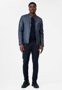 Navy Lederjacke mit tailliertem Design, ausgestattet mit silbernen Reißverschlüssen auf der Brust, gerippten Akzenten an den Schultern und einem hohen Kragen. Kombiniert mit navyfarbenen Hosen und schwarzen Sneakers.
