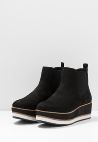 Ett par svarta mocka plattformankelstövlar med elastiska sidopaneler och lager av sula i brunt, svart och vitt på en vit yta.