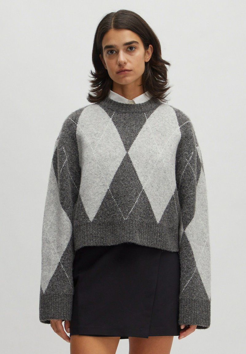 Gestrickter grauer und hellgrauer Pullover im Argyle-Muster mit weiten Ärmeln und geripptem Saum, getragen über einem weißen Hemd mit Kragen.