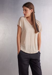 Beige top s krátkým rukávem a výstřihem do V vyrobený z lehké, texturované látky; má volný střih a jemný lesk. V kombinaci s tmavými kalhotami.