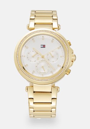 Montre-bracelet en métal doré avec cadran blanc, trois sous-cadrans, lunette sertie de cristaux et logo de marque sous la position 12 heures.