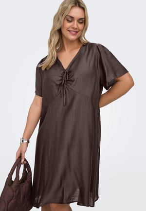 Femme souriante aux cheveux blonds ondulés porte une robe marron ample avec un col froncé, tenant un sac à main matelassé marron assorti.