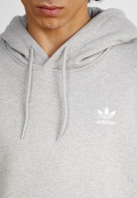 Pelēks sudraba kapuce, izgatavota no mīksta auduma, ar priekšējo kangaroo kabatu, regulējamiem auklām un baltu Adidas logo uz krūtīm.