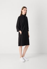 Zwart jurk met een hoge kraag en rits, voorzien van geplooide zijdetails en zakken. Glad materiaal, knie-lengte, gedragen met witte sneakers.